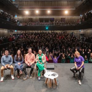 Five people are sat on a stage, with a full theatre auditorium of children behind them