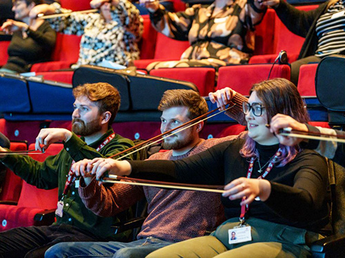 Three adults, sitting in a theatre, holding a violin bow